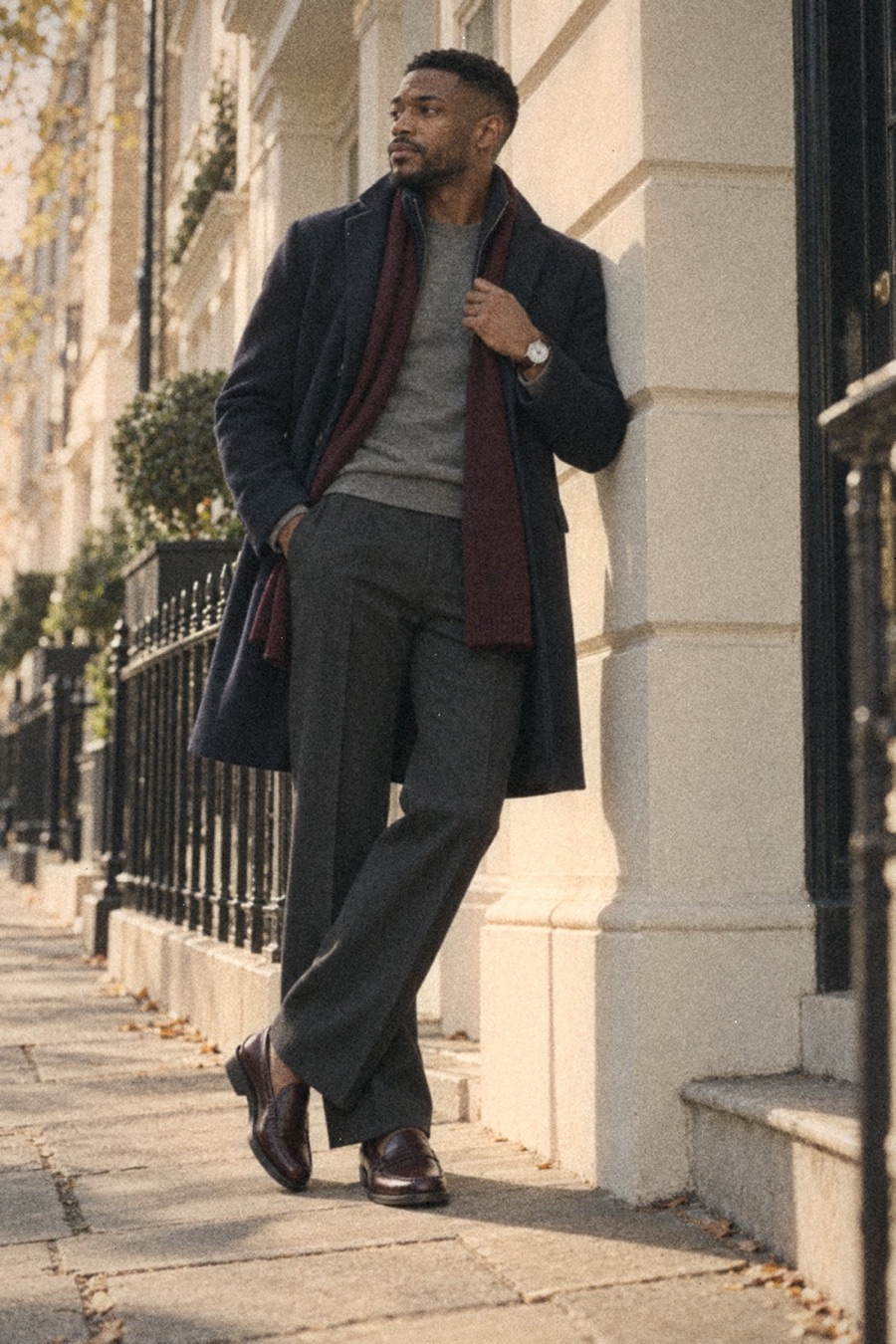 Man in a navy wool overcoat, grey knit, charcoal trousers, burgundy scarf and burgundy loafers standing against a sunlit cream stucco wall in London, looking away, silver Longines watch visible.