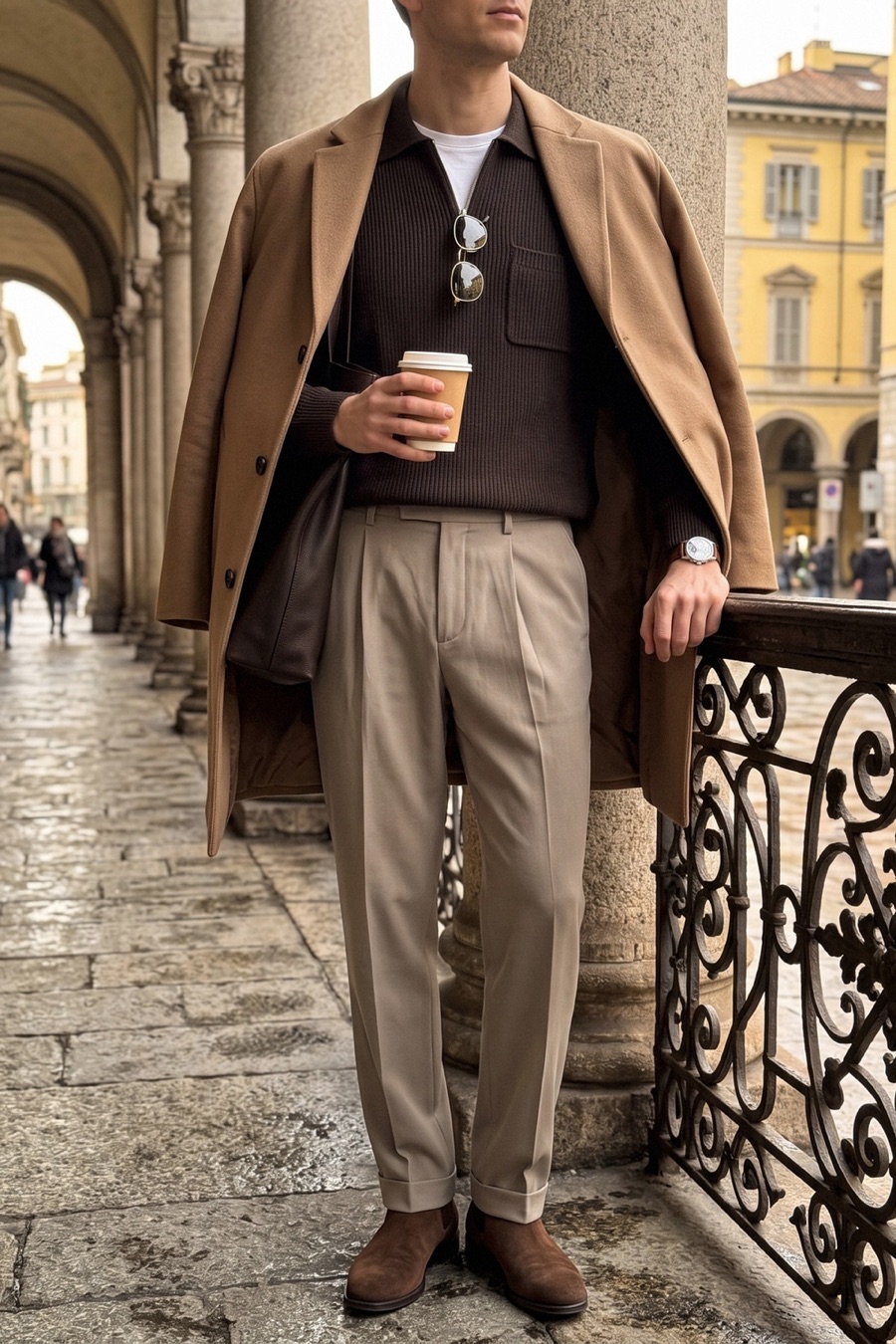 Faceless photo of a man in a camel topcoat, brown zip knit polo, mink grey pleated trousers and brown suede Chelsea boots walking beside an iron railing under a stone arcade.