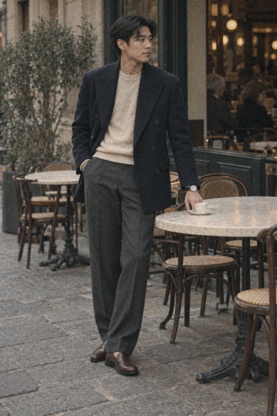 Man with black side-parted hair seated at a café table in Milan, wearing a navy double-breasted blazer over a cream cashmere sweater, charcoal trousers, brown moccasins and a blue-dial dress watch.