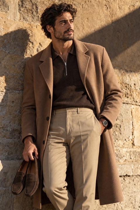 Man in a long camel wool coat, dark brown zip polo, sand trousers and dark brown suede driving loafers standing against a sunlit ochre stone wall, looking away.